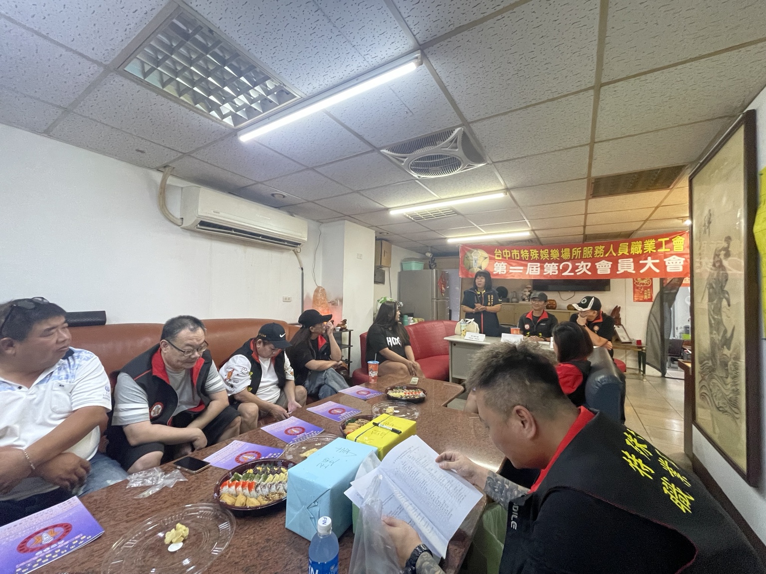 會員圍坐長桌，前方有人站立發言，後方懸掛第二屆第2次會員大會布條
